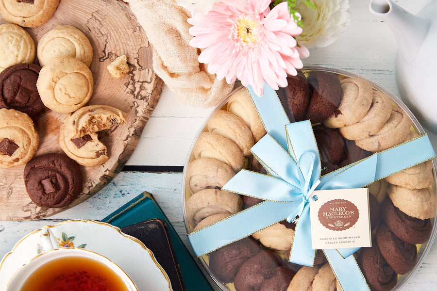 Beautiful spread of freshly baked cookies arranged on a rustic wooden board, with a cup of tea, milk jug, and a large round gift box of chocolate chunk shortbread cookies, tied with a light blue ribbon. The scene is complemented by soft flowers and a teapot, creating a cozy, inviting tea-time setting. Assorted 9