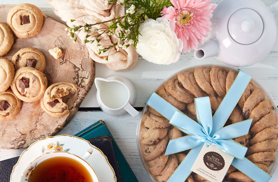 Beautiful spread of freshly baked cookies arranged on a rustic wooden board, with a cup of tea, milk jug, and a large round gift box of chocolate chunk shortbread cookies, tied with a light blue ribbon. The scene is complemented by soft flowers and a teapot, creating a cozy, inviting tea-time setting. Chocolate Crunch 9