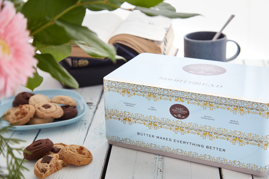 image is of a mary macleod's shortbread signature blue shortbread cookie tin on a wooden table with cookies around it. There is a cup of tea and a book in the background. This is a two (2) layer signature cookie tin.